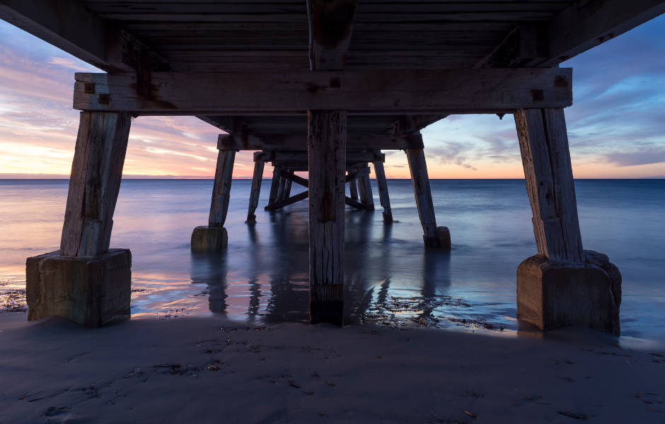 View from underneath the Normanville Jetty with a pink sunset as the backdrop.  Jetty shows wooden pylons with concrete bases in shallow water on the beach.